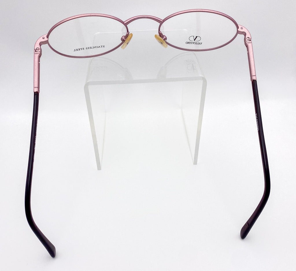Metallic lavender oval eyeglasses on a clear stand against a white background