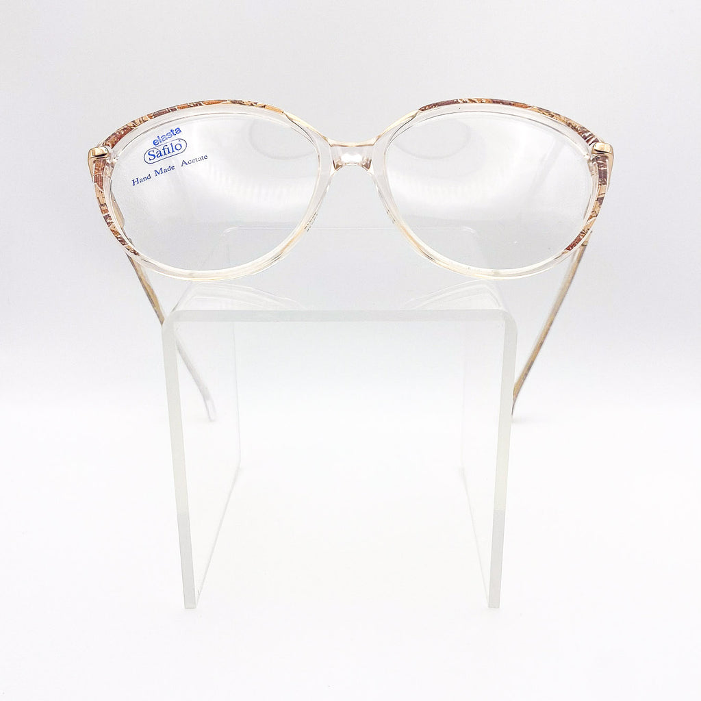 Gold-framed eyeglasses on a white stand with 'Safilo Elasta' branding.