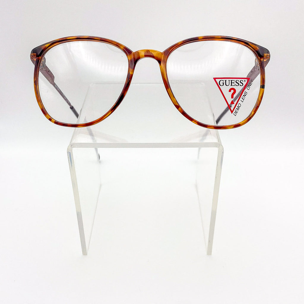 Tortoiseshell glasses with Guess logo on a clear stand against a white background