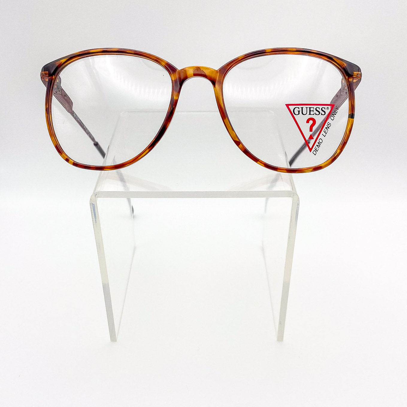 Tortoiseshell glasses with Guess logo on a clear stand against a white background