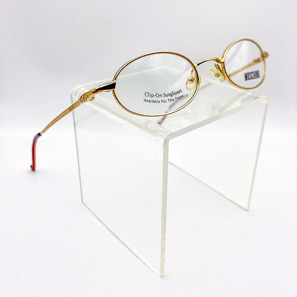 Gold-framed eyeglasses on a clear stand with 'Moschino' branding.