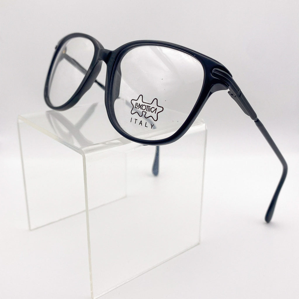 Black vintage eyeglasses with 'Luxottica Italy' logo on a clear stand against a white background
